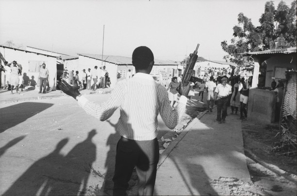 An out-of-uniform Tonton Macoute with an Uzi machine gun harasses a crowd